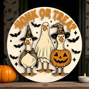 Puede incluir: Decoraci&oacute;n redonda de Halloween de madera con tres gansos disfrazados. Uno lleva un sombrero de bruja y una capa, otro es un fantasma y el tercero es una calabaza. Las palabras "HONK OR TREAT" est&aacute;n en la parte superior.