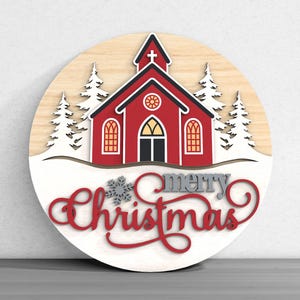 Puede incluir: Decoraci&oacute;n navide&ntilde;a redonda de madera con una iglesia roja con tejado negro y nieve blanca. Las palabras "merry Christmas" est&aacute;n escritas en escritura roja y gris. Los &aacute;rboles blancos flanquean la iglesia.