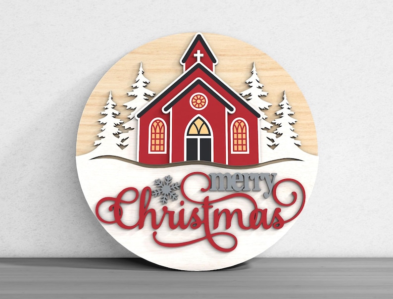 Puede incluir: Decoraci&oacute;n navide&ntilde;a redonda de madera con una iglesia roja con tejado negro y nieve blanca. Las palabras "merry Christmas" est&aacute;n escritas en escritura roja y gris. Los &aacute;rboles blancos flanquean la iglesia.