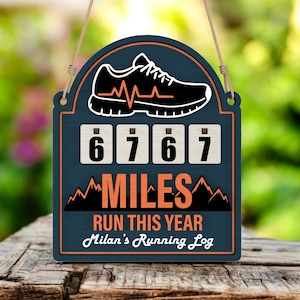 Op de afbeelding: Houten bord met een hardloopschoenafbeelding en de tekst "6767 MILES RUN THIS YEAR Milan's Running Log". Het bord is donkerblauw met oranje en witte accenten en een beige touw om op te hangen.
