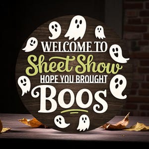 Puede incluir: Un letrero redondo de madera de Halloween con el texto "WELCOME TO Sheet Show HOPE YOU BROUGHT BOOS" en letras blancas y verdes. Recortes de fantasmas blancos rodean el texto. El letrero est&aacute; sobre una superficie de madera con hojas secas.