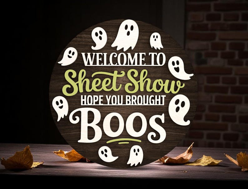 Puede incluir: Un letrero redondo de madera de Halloween con el texto "WELCOME TO Sheet Show HOPE YOU BROUGHT BOOS" en letras blancas y verdes. Recortes de fantasmas blancos rodean el texto. El letrero est&aacute; sobre una superficie de madera con hojas secas.