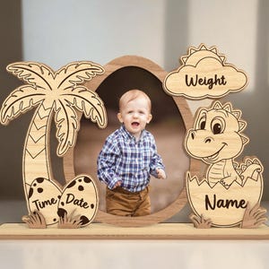 Può includere: Cornice per foto in legno con un dinosauro, una palma e uova. La cornice ha spazi per una foto, un nome, un peso, un'ora e una data.