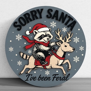 May include: A round, gray wooden sign with a raccoon riding a reindeer, both wearing Christmas attire. The sign reads "Sorry Santa, I've been Feral" in black lettering, with snowflakes scattered around the image.
