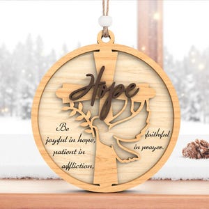 Può includere: Ornamento natalizio in legno con croce, colomba e la parola "Hope". L'ornamento include il testo "Be joyful in hope, patient in affliction, faithful in prayer."