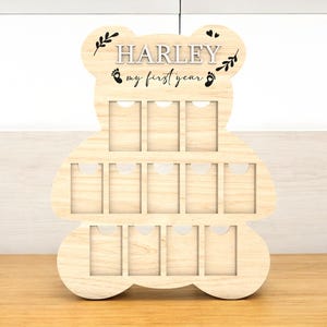 Può includere: Un espositore fotografico in legno a forma di orso con il nome "HARLEY" e il testo "my first year". L'espositore ha più fessure rettangolari per le foto ed è realizzato in legno chiaro. Decorato con foglie e impronte.