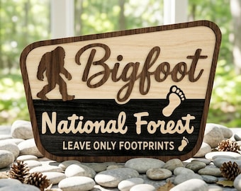 Puede incluir: Un letrero de madera trapezoidal con una sección superior de madera clara que presenta una silueta de Bigfoot marrón oscuro y el texto "Bigfoot" en cursiva. La sección inferior es de madera oscura con el texto "National Forest" y "LEAVE ONLY FOOTPRINTS".