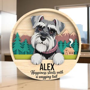 May include: A round wooden sign featuring a Schnauzer dog design. The sign includes the name "ALEX" and the phrase "Happiness starts with a wagging tail." The background depicts mountains, trees, and a house.