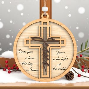 May include: A wooden Christmas ornament featuring a cross design with the word "Jesus" and the phrases "Unto you is born this day a Savior" and "Jesus is the light of the world." The ornament is round with a light brown finish.