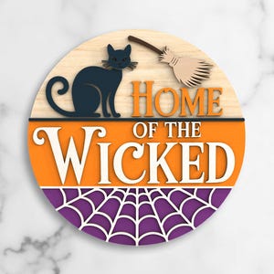 May include: A circular Halloween sign featuring a black cat, a broom, and the phrase "HOME OF THE WICKED." The sign is made of wood and has orange, purple, and cream tones. A spider web design is at the bottom.
