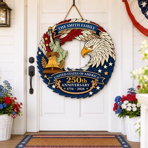 May include: A wooden wreath with a patriotic theme, featuring an eagle, the Statue of Liberty, and the Liberty Bell. The wreath is adorned with red, white, and blue elements and includes the text "United States of America 250th Anniversary 1776-2026."