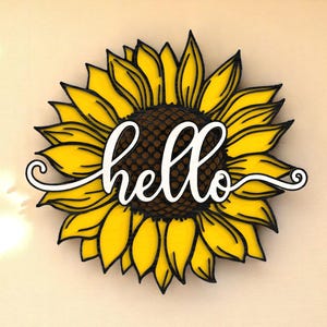 May include: A wooden sunflower decoration with yellow petals, a brown center, and the word "hello" in white script. The sunflower has black outlines and is set against a neutral background.