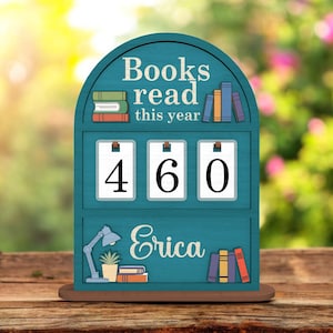Op de afbeelding: Een turquoise houten boekensteller met de tekst "Boeken gelezen dit jaar" en het nummer "460". De naam "Erica" staat eronder. Het ontwerp bevat illustraties van boeken, een bureaulamp en een potplant.