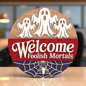 May include: A circular wooden Halloween sign featuring three white ghost cutouts above the words "Welcome Foolish Mortals". The sign has red and blue sections with a spider web and spider design.