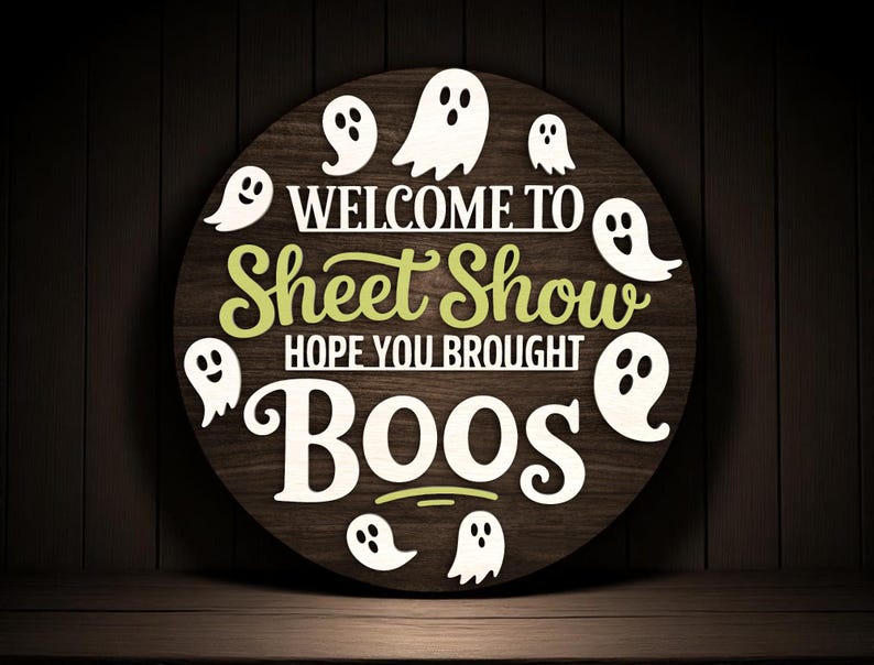 Puede incluir: Un letrero redondo de madera con el texto "WELCOME TO Sheet Show HOPE YOU BROUGHT BOOS" en letras blancas y verdes. Siluetas de fantasmas blancos rodean el texto. El letrero est&aacute; sobre un fondo de madera oscura, ideal para Halloween.
