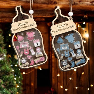 Op de afbeelding: Twee houten kerstversieringen in de vorm van een babyfles. Eén is roze met "Ellie's First Christmas 2025" en de andere is blauw met "Liam's First Christmas 2025". Elke versiering heeft baby-gerelateerde iconen en de tijd 09:30 uur.