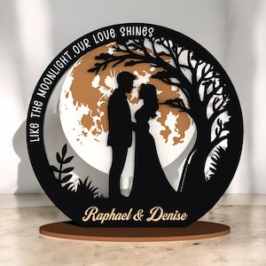 May include: A black wooden silhouette of a couple under a tree, with a moon backdrop. The text "LIKE THE MOONLIGHT, OUR LOVE SHINES" is above the couple, and "Raphael & Denise" is below. The base is brown.