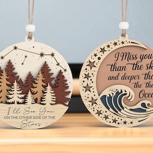 May include: Two wooden round ornaments with twine hangers. One features a forest scene with the text "I'll See You On The Other Side Of The Stars." The other has a moon, stars, and ocean wave design with the text "I Miss You Than The Sky And Deeper Than The Ocean."