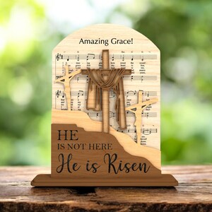 May include: Wooden religious decoration featuring three crosses, with the central cross draped with fabric. The background includes sheet music and the words "Amazing Grace!" and "He is not here, He is Risen."