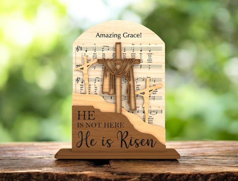 May include: Wooden religious decoration featuring three crosses, with the central cross draped with fabric. The background includes sheet music and the words "Amazing Grace!" and "He is not here, He is Risen."