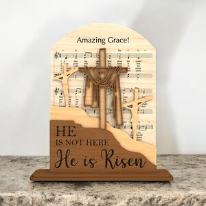 May include: Wooden religious decoration featuring three crosses and the text "He is not here, He is Risen." The background includes sheet music and the words "Amazing Grace!" in a decorative wooden frame.