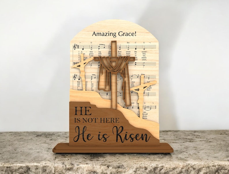May include: Wooden religious decoration featuring three crosses and the text "He is not here, He is Risen." The background includes sheet music and the words "Amazing Grace!" in a decorative wooden frame.