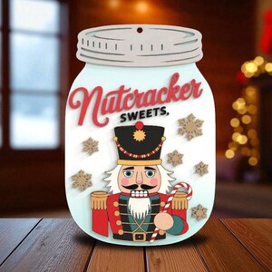 May include: A wooden decoration shaped like a mason jar with a light blue background. The jar features the words "Nutcracker Sweets" in red script, a nutcracker soldier, and snowflake accents. The nutcracker is wearing a black hat and red jacket.