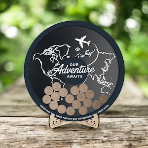 May include: A round, black travel-themed coin bank with a world map design and the words "Our Adventure Awaits." A white airplane graphic is above the text. The bank is filled with coins and sits on a wooden stand. The text "Coins may fill your pocket but adventure will your soul" is at the bottom.