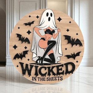 May include: Circular wooden sign featuring a pin-up ghost illustration. The ghost is wearing lingerie and thigh-high boots. Black bats and diamond shapes surround the image. The text "WICKED IN THE SHEETS" is at the bottom.