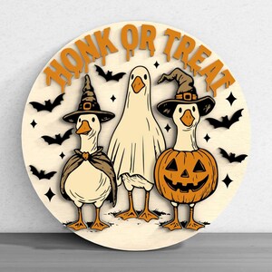 Puede incluir: Decoraci&oacute;n redonda de Halloween de madera con tres gansos disfrazados. Uno es un fantasma, otro una bruja y otro una calabaza. Las palabras "HONK OR TREAT" est&aacute;n en la parte superior, con murci&eacute;lagos y estrellas alrededor de los gansos.