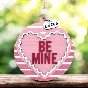 May include: A pink and white Valentine's Day ornament with the words "BE MINE" in red. The ornament has a heart shape with a scalloped edge and a small tag that says "Lucas". It hangs from a natural twine.