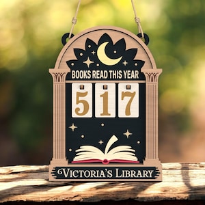 Könnte beinhalten: Holzschild mit dem Text "BOOKS READ THIS YEAR" und der Zahl "517" auf schwarzem Hintergrund. Das Schild zeigt auch einen Halbmond, Sterne und ein offenes Buch. Auf der Unterseite des Schildes steht "Victoria's Library".