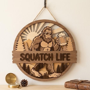 May include: A circular wooden wall hanging featuring a layered design of a Bigfoot character wearing sunglasses and holding a beer mug. The text SQUATCH LIFE is carved across the centre. The scene includes pine trees and a sunburst background.