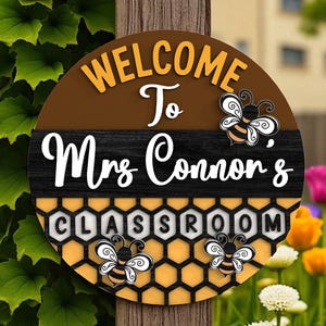 Peut inclure: Panneau rond en bois avec le texte "WELCOME To Mrs Connors CLASSROOM". Le panneau présente un dessus marron, une section centrale noire et un fond en nid d'abeille. Des motifs d'abeilles décoratives sont inclus.