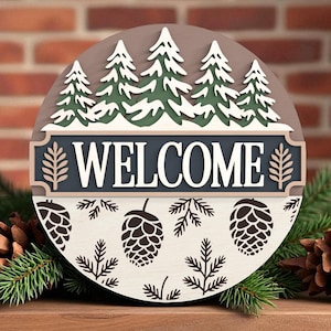 Könnte beinhalten: Ein rundes Holzschild mit Waldmotiv. Das Schild zeigt das Wort "WELCOME" in weißen Buchstaben auf dunkelblauem Hintergrund, mit Kiefern und Tannenzapfen. Das Schild ist von Tannennadeln und Tannenzapfen umgeben.