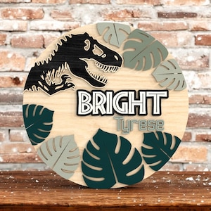 Può includere: Un cartello rotondo in legno con un dinosauro nero e il nome "Bright Tyrese". Il cartello presenta foglie di monstera verdi e grigie e il nome in grassetto. Lo sfondo è di colore legno chiaro.