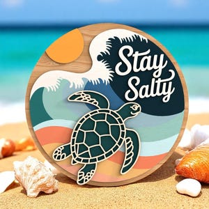 May include: A round wooden sign with a beach theme. It features a sea turtle, waves, and the text "Stay Salty" in white. The background shows a beach with seashells and the ocean.