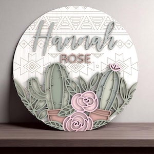 Könnte beinhalten: Runder Holzschild mit dem Namen "Hannah Rose" in 3D-Schrift. Das Schild zeigt ein Kaktus- und Blumendesign in Grün- und Rosatönen, vor einem weißen Hintergrund mit einem geometrischen Muster.