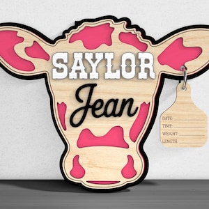 May include: Wooden cow head-shaped wall decor with the name "Saylor Jean" in white and black lettering. The cow has pink spots and a black outline. A small tag with lines for date, time, weight, and length hangs from the ear.