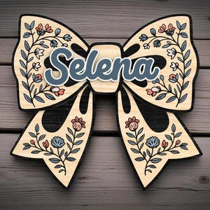 Puede incluir: Decoración de pared de madera con forma de lazo con el nombre "Selena" en escritura azul. El lazo es de madera clara con bordes negros y diseños florales en azul, rosa y blanco. Adecuado para un dormitorio o guardería.