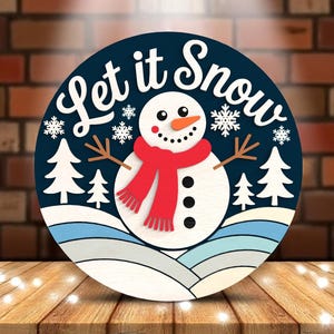 Könnte beinhalten: Runder Holzschild mit einem Schneemann-Design. Der Schneemann hat einen roten Schal und eine Karottennase. Auf dem Schild steht "Let it Snow" in weißer Schrift, mit Schneeflocken und Tannenbäumen.