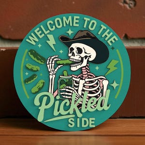 Pode incluir: Adesivo redondo turquesa com um esqueleto usando um chapéu de cowboy, comendo um picles. O texto "WELCOME TO THE PICKLED SIDE" está em verde claro, com raios e estrelas.