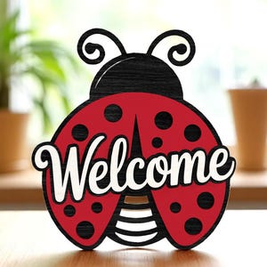 Może przedstawiać: Drewniany znak w kształcie biedronki z napisem "Welcome" w białym skrypcie. Biedronka ma czarną głowę i czerwone skrzydła z czarnymi kropkami. Dekoracyjny przedmiot do domu.