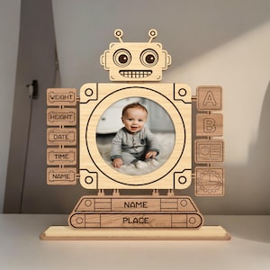 Puede incluir: Marco de fotos de robot de madera con un recorte circular para una foto. El marco tiene un acabado de madera y presenta una cara de robot con ojos grandes y una sonrisa. El marco tiene ranuras para etiquetas con las palabras "Peso", "Altura", "Fecha", "Hora", "Nombre", "Lugar", "A", "B" y "C".