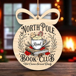 May include: Wooden Christmas bauble with the text "North Pole Book Club" and a graphic of a mug of hot cocoa, a gingerbread man, and a stack of books. The bauble is round with a bow-shaped top and a string for hanging.