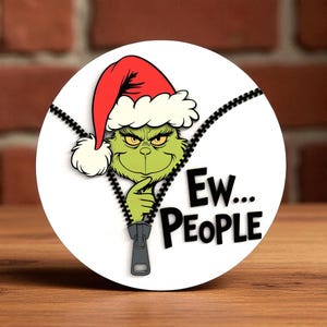 Puede incluir: Un diseño redondo y blanco con el Grinch con un gorro de Papá Noel y el texto "Ew... PEOPLE" en negro. El Grinch es verde con ojos amarillos y aparece detrás de una cremallera negra.