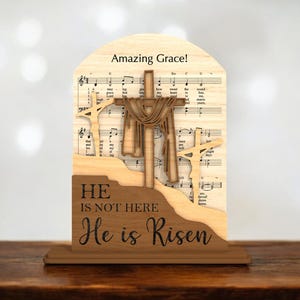 May include: Wooden Easter decoration featuring three crosses, with the central cross draped. The background includes sheet music and the words "Amazing Grace!" and "He is not here, He is Risen."