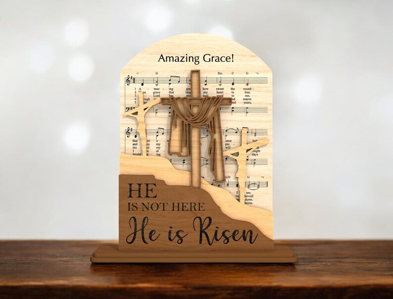 May include: Wooden Easter decoration featuring three crosses, with the central cross draped. The background includes sheet music and the words "Amazing Grace!" and "He is not here, He is Risen."