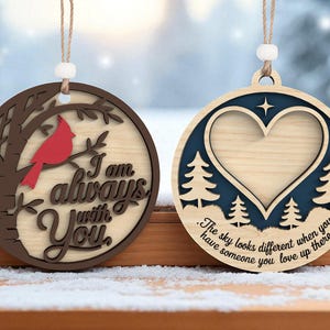 May include: Two wooden Christmas ornaments. One features a red cardinal on a tree branch with the text "I am always with You." The other has a heart shape, trees, and the text "The sky looks different when you have someone you love up there."