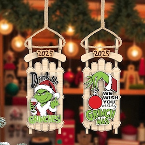 Puede incluir: Dos adornos navideños de madera con forma de trineo. Uno presenta al Grinch con una copa de vino y el texto "Drink Up Grinches!". El otro dice "We Wish You Merry Grinchmas!" con una mano del Grinch.